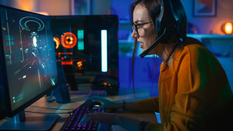 Pretty and Excited Gamer Girl in Glasses and Headset with a Mic Playing Shooter Online Video Game on Her Computer. Room and PC have Colorful Neon Led Lights. Cozy Evening at Home.