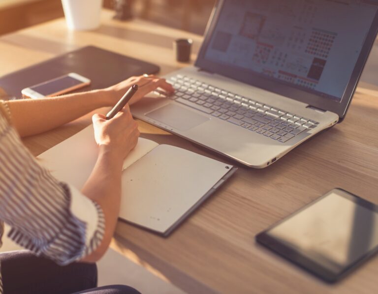 Female designer using laptop, sketching at blank notepad. Woman hand writing in notebook on wooden desk.