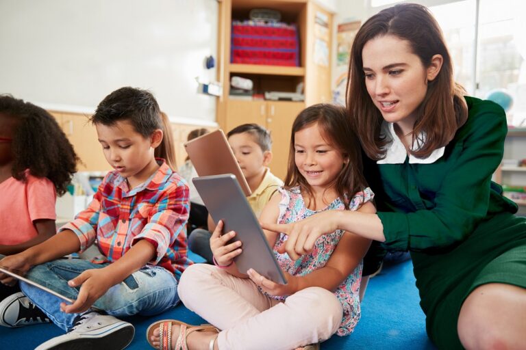 Teacher,Helps,Girl,In,Elementary,Class,With,Tablet,Computers